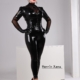Domina Herrin Xena steht in schwarzem Latexanzug mit Korsett und transparenten Ärmeln, die Hände an den Hüften, vor hellem Studiohintergrund.