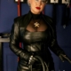 Domina Herrin Xena steht in schwarzer Lederjacke, Lederkorsettkleid und Lederhandschuhen mit einer Peitsche vor einer Wand mit Schlaginstrumenten.