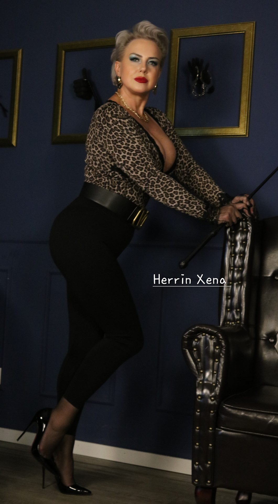 Domina Herrin Xena steht in Leopardenoberteil, breitem Gürtel und schwarzer Hose mit einer Gerte neben einem schwarzen Ledersessel vor dunkelblauer Wand.