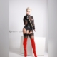 Domina Herrin Xena steht in transparentem Body mit Latexkorsett, Strapsen, schwarzen Strümpfen und roten Lack-Overknees und hält einen Rohrstock.