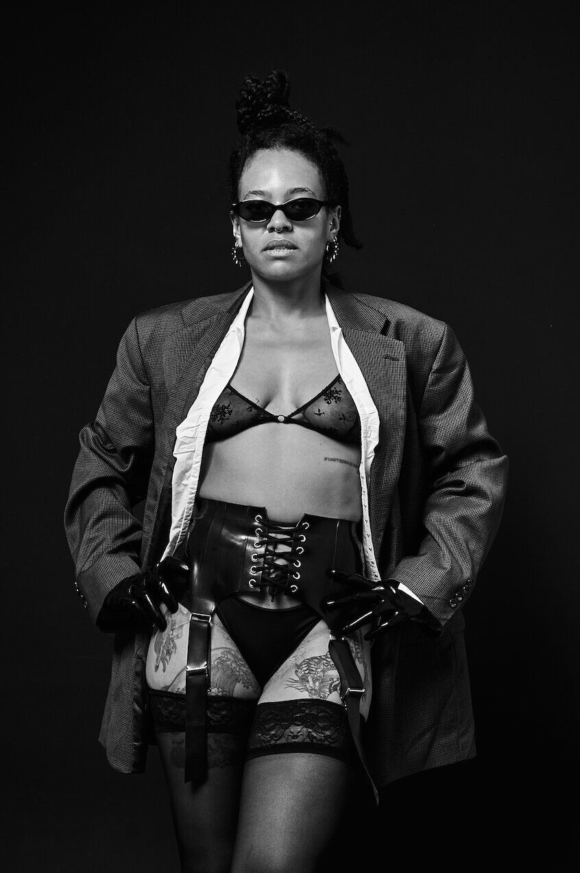 Bizarrlady Kiki Reyna mit Sonnenbrille, offenem Blazer, Dessous, Korsettgürtel und Strümpfen vor schwarzem Hintergrund, Schwarzweissaufnahme.