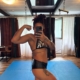 Bizarrlady Kiki Reyna fotografiert sich im Spiegel in Bikini-Top und Shorts auf blauen Bodenmatten und zeigt ihren Bizeps.