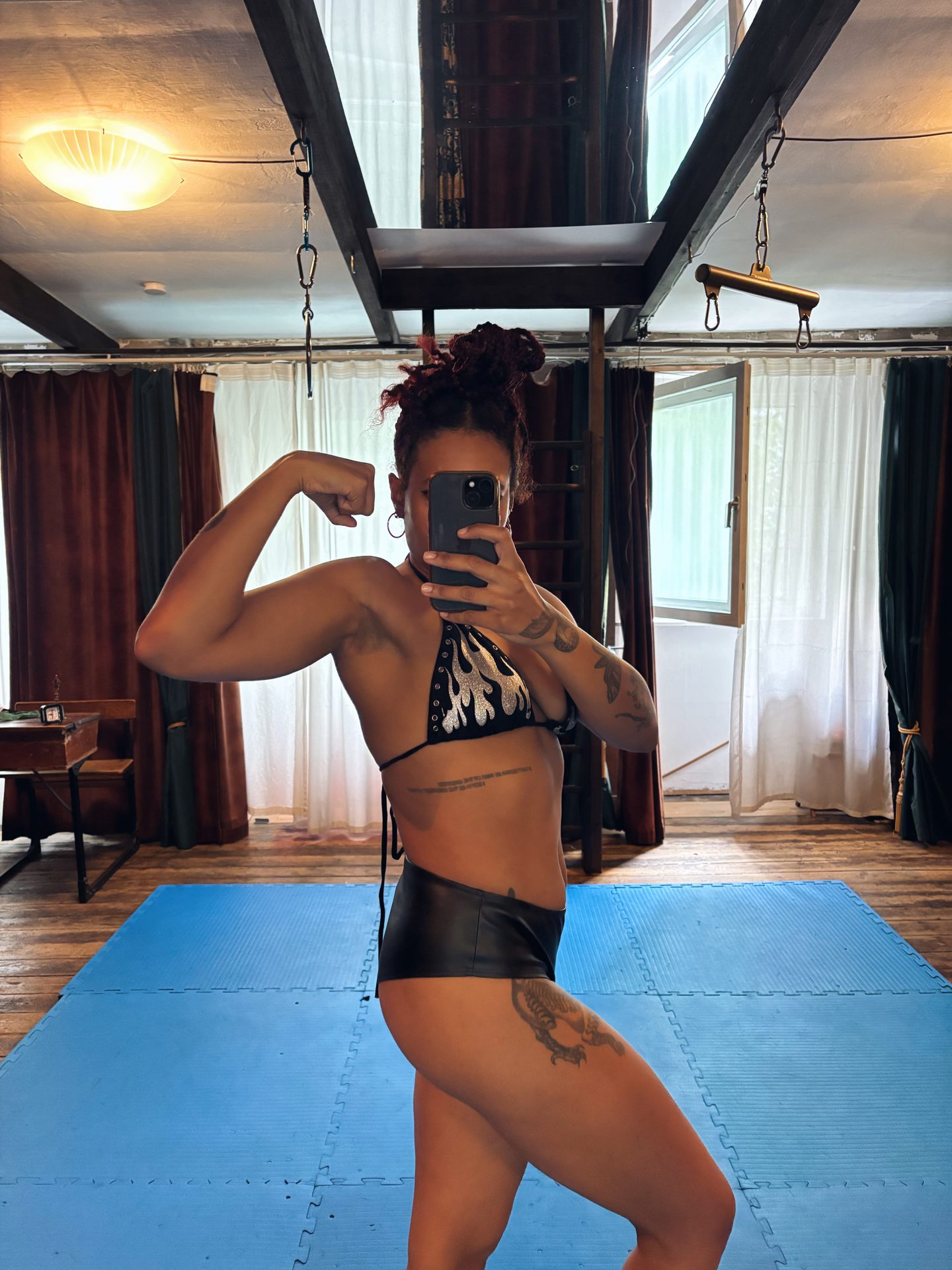 Bizarrlady Kiki Reyna fotografiert sich im Spiegel in Bikini-Top und Shorts auf blauen Bodenmatten und zeigt ihren Bizeps.
