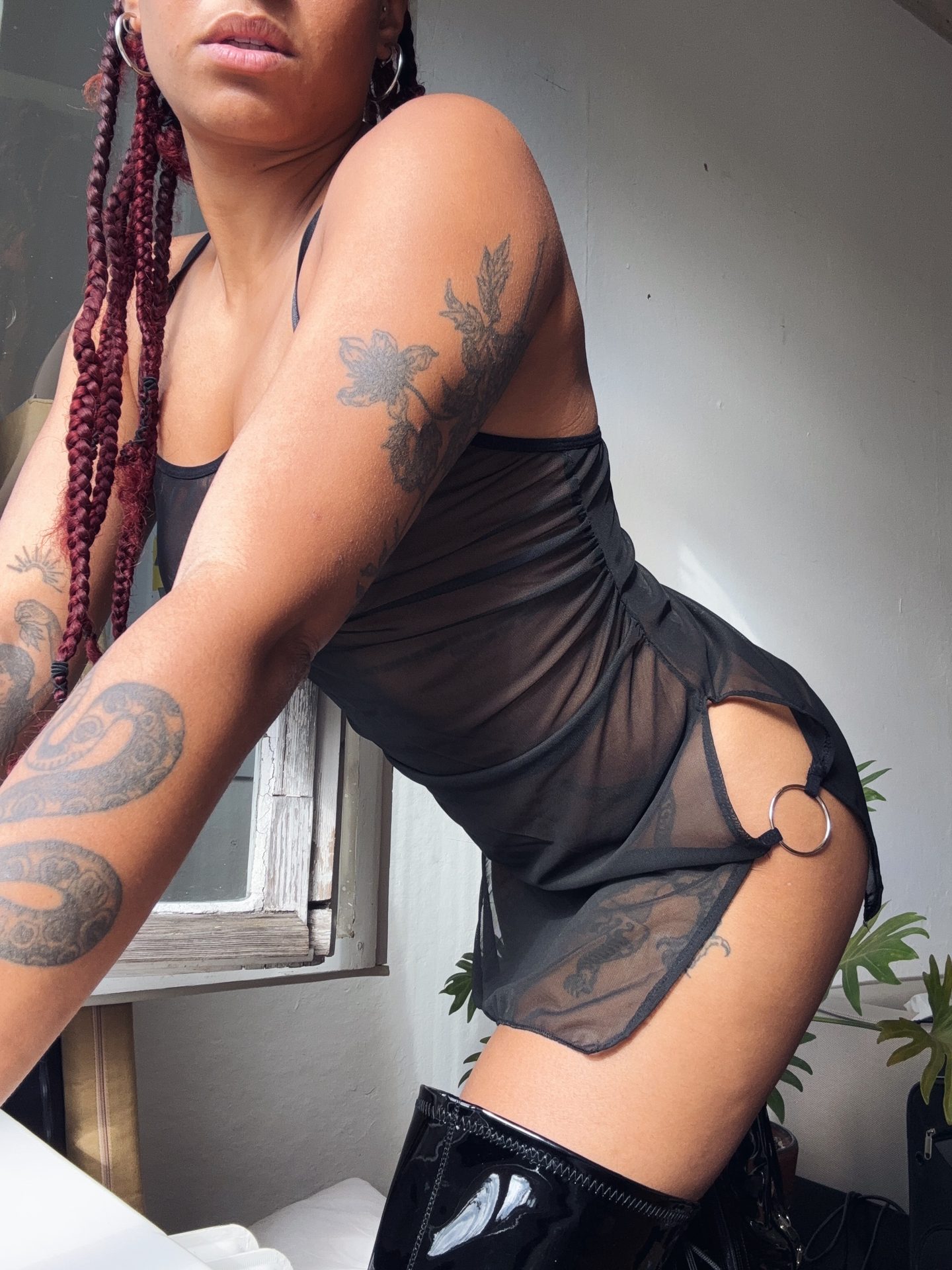 Bizarrlady Kiki Reyna mit langen roten Zöpfen und Tattoos im transparenten schwarzen Kleid, leicht vorgebeugt.