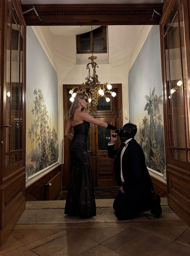 Lady Diyosa im Abendkleid unter Kronleuchter Domina Lady Diyosa steht im langen schwarzen Kleid unter einem Kronleuchter; neben ihr kniet eine maskierte Person im Anzug.