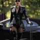 Lady Diyosa mit Rollkoffer vor Cabrio Domina Lady Diyosa mit Sonnenbrille und schwarzem Blazer, eine Hand am Rollkoffer, vor einem schwarzen Cabrio.