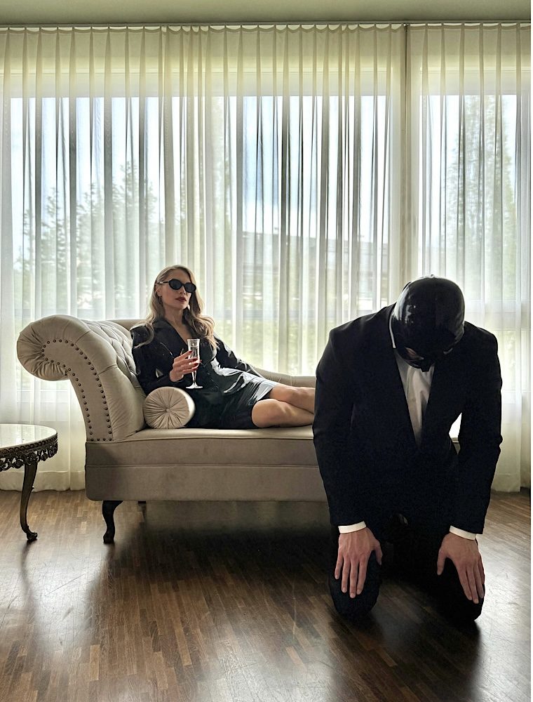 Lady Diyosa auf Chaiselongue mit kniender Person Domina Lady Diyosa sitzt mit einem Glas auf einer hellen Chaiselongue; davor kniet eine maskierte Person im schwarzen Anzug.