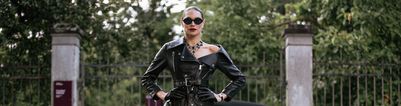 Domina Lady Diyosa mit Sonnenbrille und schwarzer Lederjacke vor einem Eisentor mit Steinpfeilern.