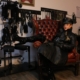 Domina Lady Lia sitzt mit Schirmmütze und Ledermantel in einem roten Chesterfield-Sessel vor einer Wand mit Schlaginstrumenten und Fesseln.