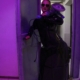 Domina Lady Lia mit Sonnenbrille und langem schwarzem Lackmantel steht im violetten Licht in einer offenen Stahltür und hält ein Elektrowerkzeug.