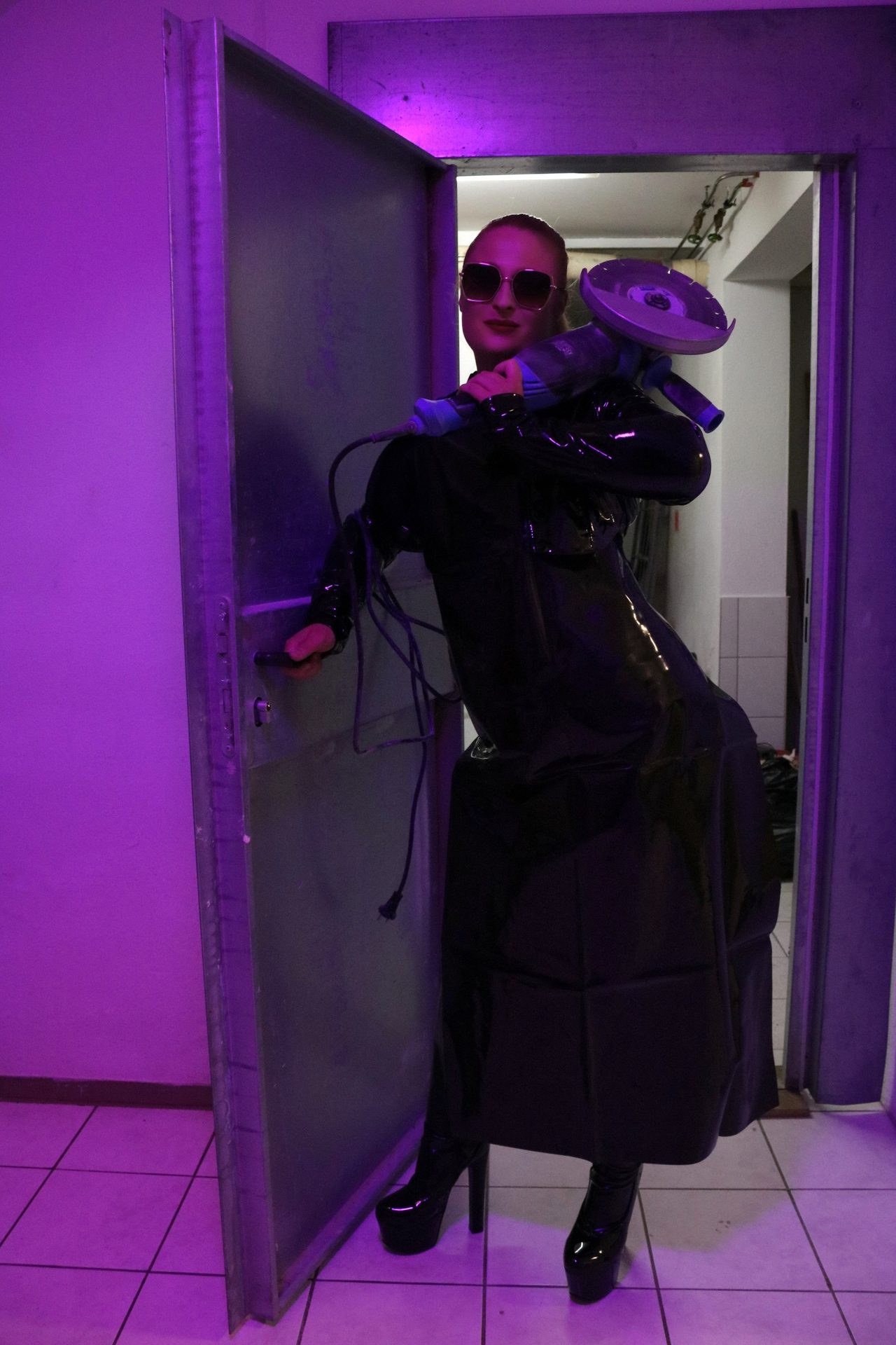Domina Lady Lia mit Sonnenbrille und langem schwarzem Lackmantel steht im violetten Licht in einer offenen Stahltür und hält ein Elektrowerkzeug.